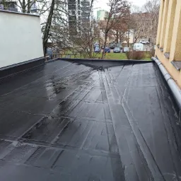 Czarna, płaska powierzchnia dachu pokryta papą termozgrzewalną, z widocznymi łączeniami i odbiciami światła, obok ściana budynku z rurą spustową.