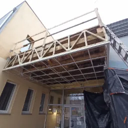 Montaż drewnianej konstrukcji zadaszenia nad wejściem do budynku biblioteki publicznej, widoczna drabina oparta o konstrukcję, zabezpieczenie folią budowlaną.