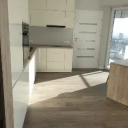 Jasna, nowoczesna kuchnia z białymi szafkami i drewnianą podłogą, widok na balkon i panoramę miasta przez duże okno.