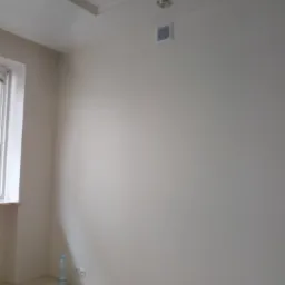 Pomalowane na jasno ściany w pustym pokoju z widoczną butelką wody mineralnej przy gniazdku elektrycznym, sufit z oświetleniem punktowym i kratką wentylacyjną.