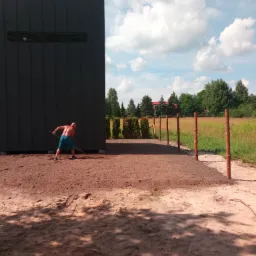 Mężczyzna wyrównuje ziemię grabiami przed nowoczesnym budynkiem z ciemną elewacją, widoczne drewniane słupki ogrodzenia i zielone drzewa w tle.