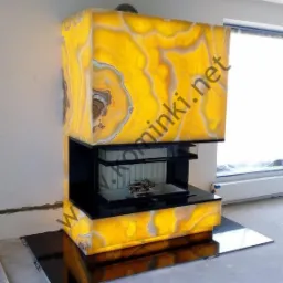 Nowoczesny kominek obudowany podświetlanym, żółtym onyksem z widocznymi naturalnymi wzorami, czarny wkład kominkowy z ułożonym drewnem wewnątrz, na czarnej, błyszczącej płycie podłogowej.