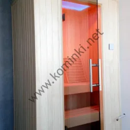 Jasna, drewniana kabina z otwartymi szklanymi drzwiami, ukazująca wnętrze z ławkami i oświetleniem w odcieniach pomarańczy.