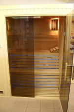 Wnętrze sauny z jasnego drewna z przeszklonymi drzwiami przesuwnymi. Widoczne drewniane ławki, oświetlenie punktowe i fragment półek z akcesoriami w tle.