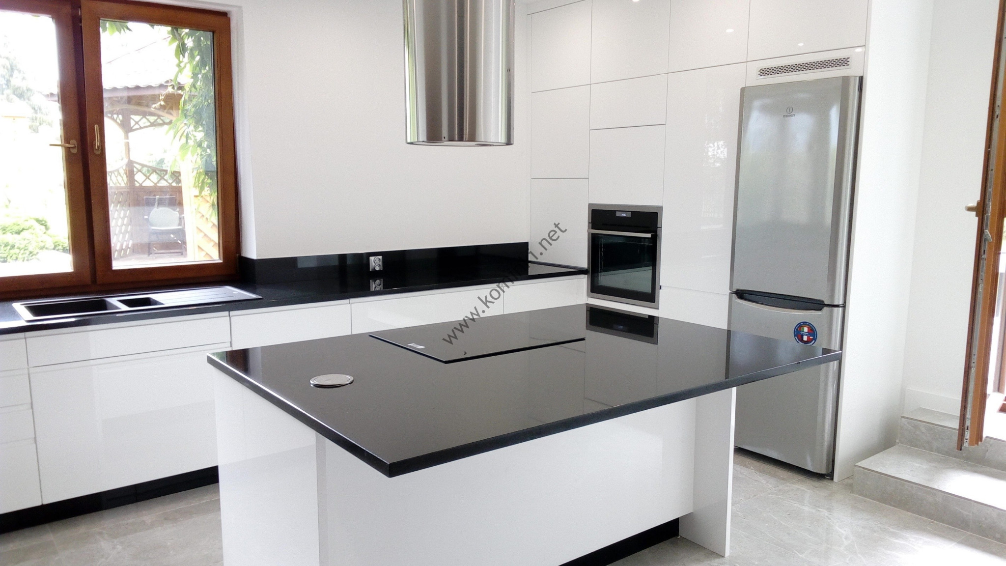 Nowoczesna kuchnia z białymi szafkami, czarnym blatem i wyspą kuchenną z płytą indukcyjną, widok na okno i ogród.