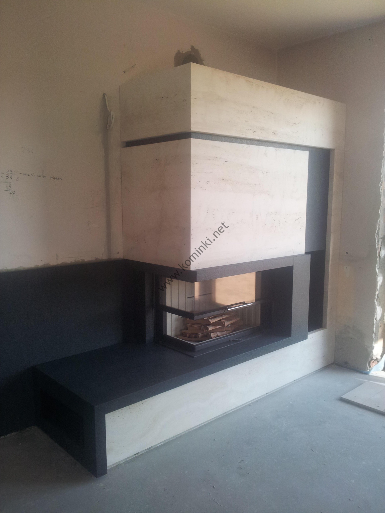 Nowoczesny kominek z obudową z jasnego trawertynu i czarnymi elementami, widoczne ułożone drewno w palenisku, ściany w trakcie wykończenia.