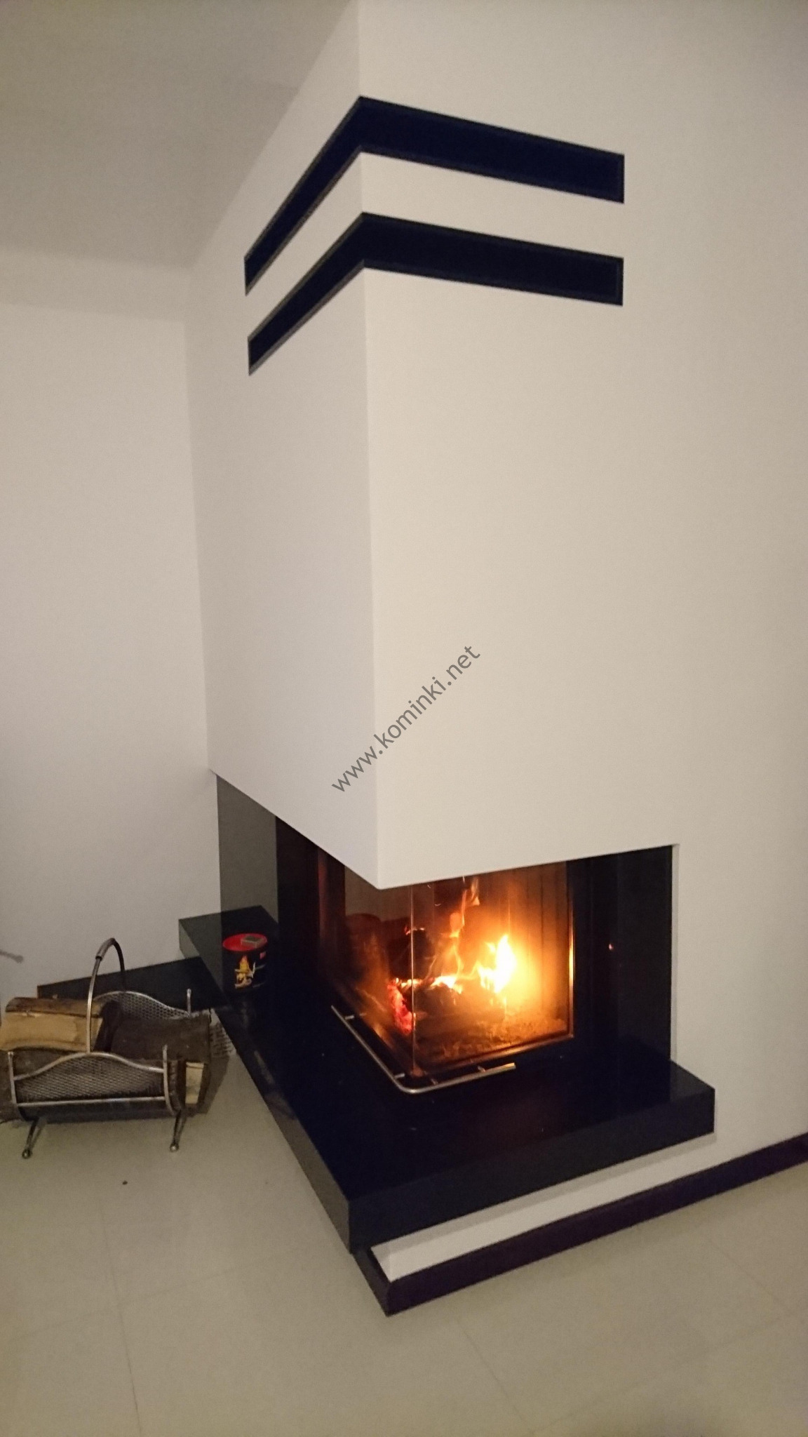 Nowoczesny kominek narożny z czarnymi akcentami, płomienie widoczne przez szklaną szybę, obok kosz z drewnem i puszka, białe ściany z ciemnymi pasami u góry.