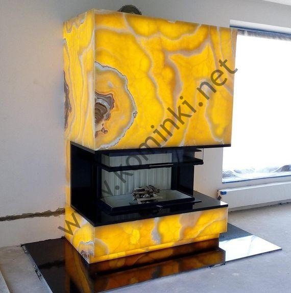 Nowoczesny kominek obudowany podświetlanym, żółtym onyksem z widocznymi naturalnymi wzorami, czarny wkład kominkowy z ułożonym drewnem wewnątrz, na czarnej, błyszczącej płycie podłogowej.