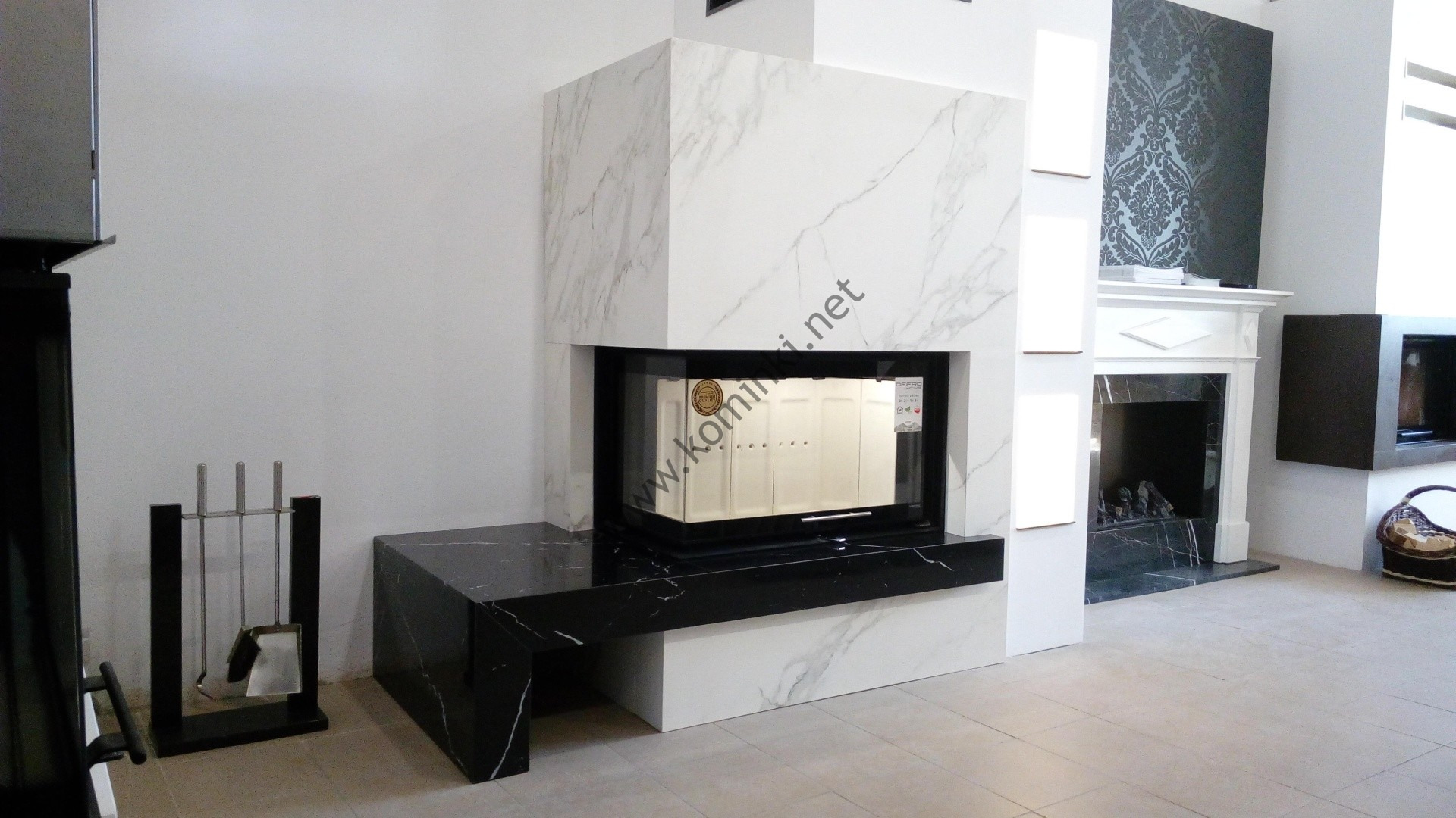 Nowoczesny kominek z obudową z białego marmuru z czarnym blatem, widoczne akcesoria kominkowe i inny kominek w tle.