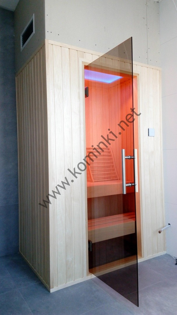 Jasna, drewniana kabina z otwartymi szklanymi drzwiami, ukazująca wnętrze z ławkami i oświetleniem w odcieniach pomarańczy.