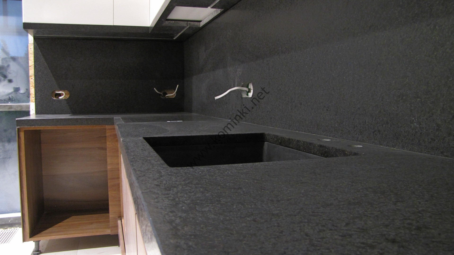 Czarny, granitowy blat kuchenny z wbudowanym zlewem, widoczne gniazdka elektryczne i drewniane szafki pod spodem.