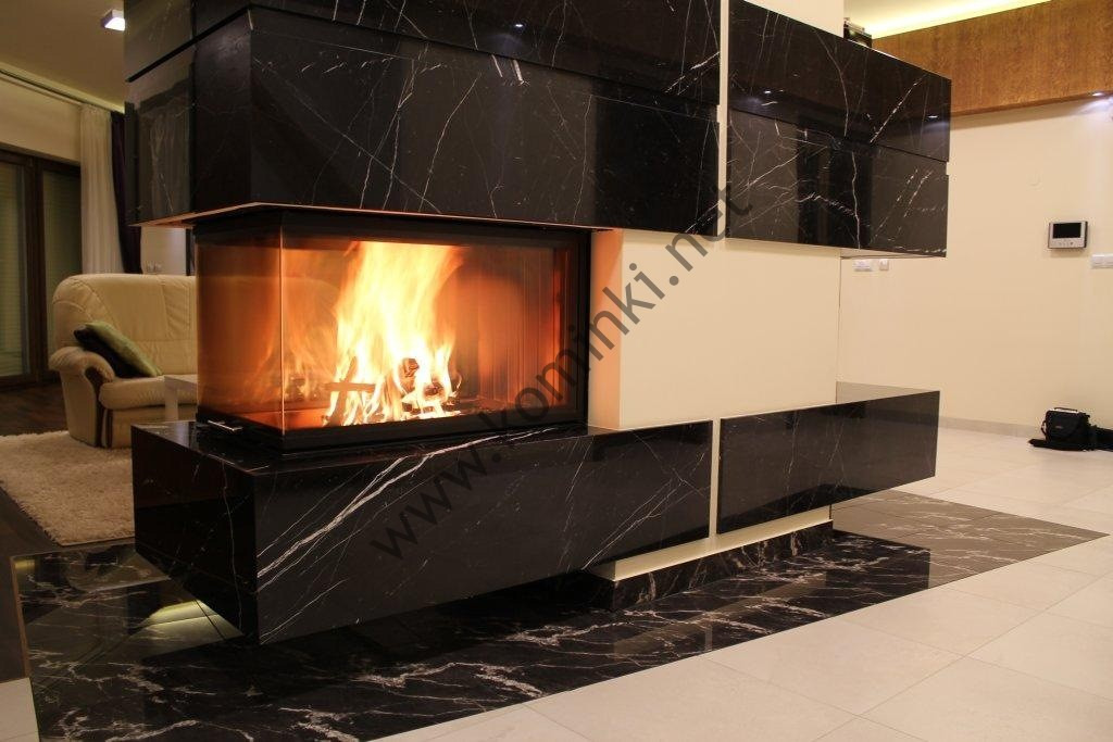 Nowoczesny kominek z czarnego marmuru z widocznym płomieniem, osadzony w minimalistycznym wnętrzu z jasnymi ścianami i podłogą.