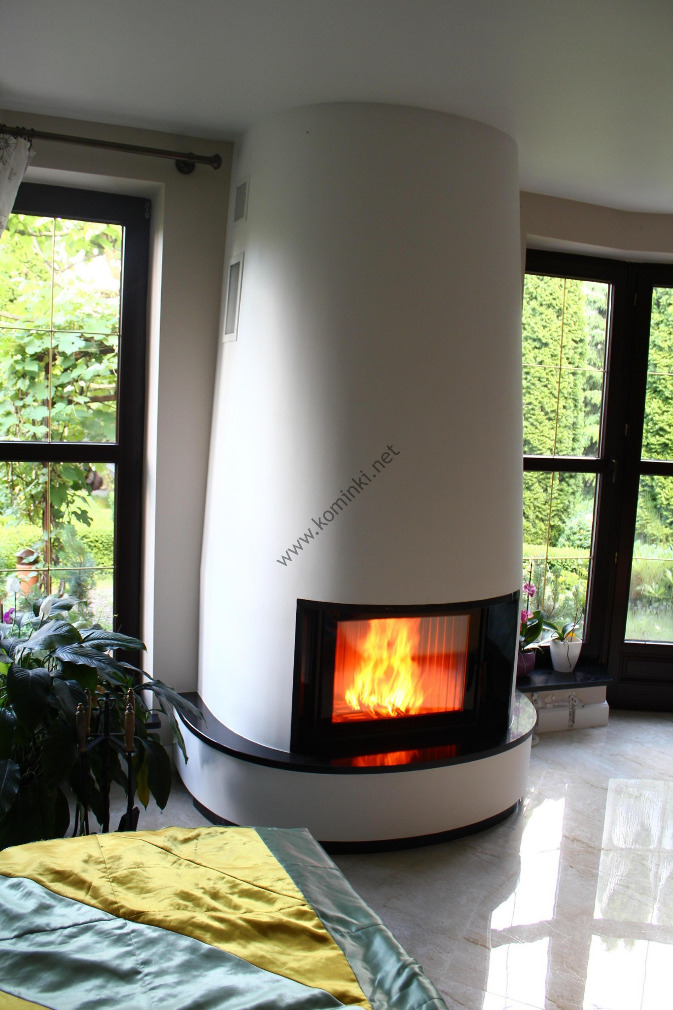 Nowoczesny, okrągły kominek w białej obudowie z płomieniami widocznymi przez czarną szybę, otoczony roślinami doniczkowymi i z widokiem na ogród przez okno.