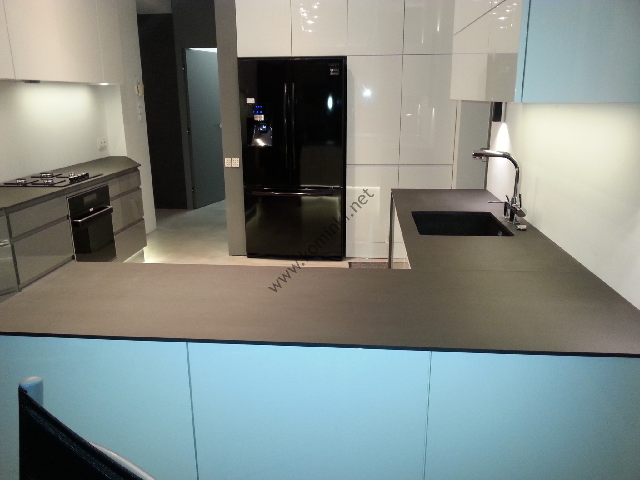 Nowoczesna kuchnia z czarnym blatem, białymi szafkami i czarną lodówką Samsung z dystrybutorem wody, widoczny zlew ze srebrną baterią i płyta grzewcza w tle.