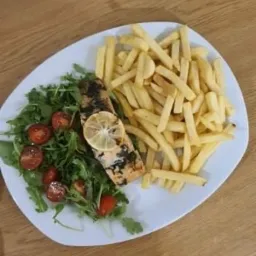 Porcja grillowanego łososia z plasterkiem cytryny, podana z sałatką z rukoli i pomidorków koktajlowych oraz frytkami na białym talerzu, widoczne drewniane tło.