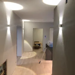 Korytarz w trakcie remontu z zamontowanymi kinkietami dającymi podwójne światło skierowane ku górze i dołowi, widoczne kartony w głębi pomieszczenia.