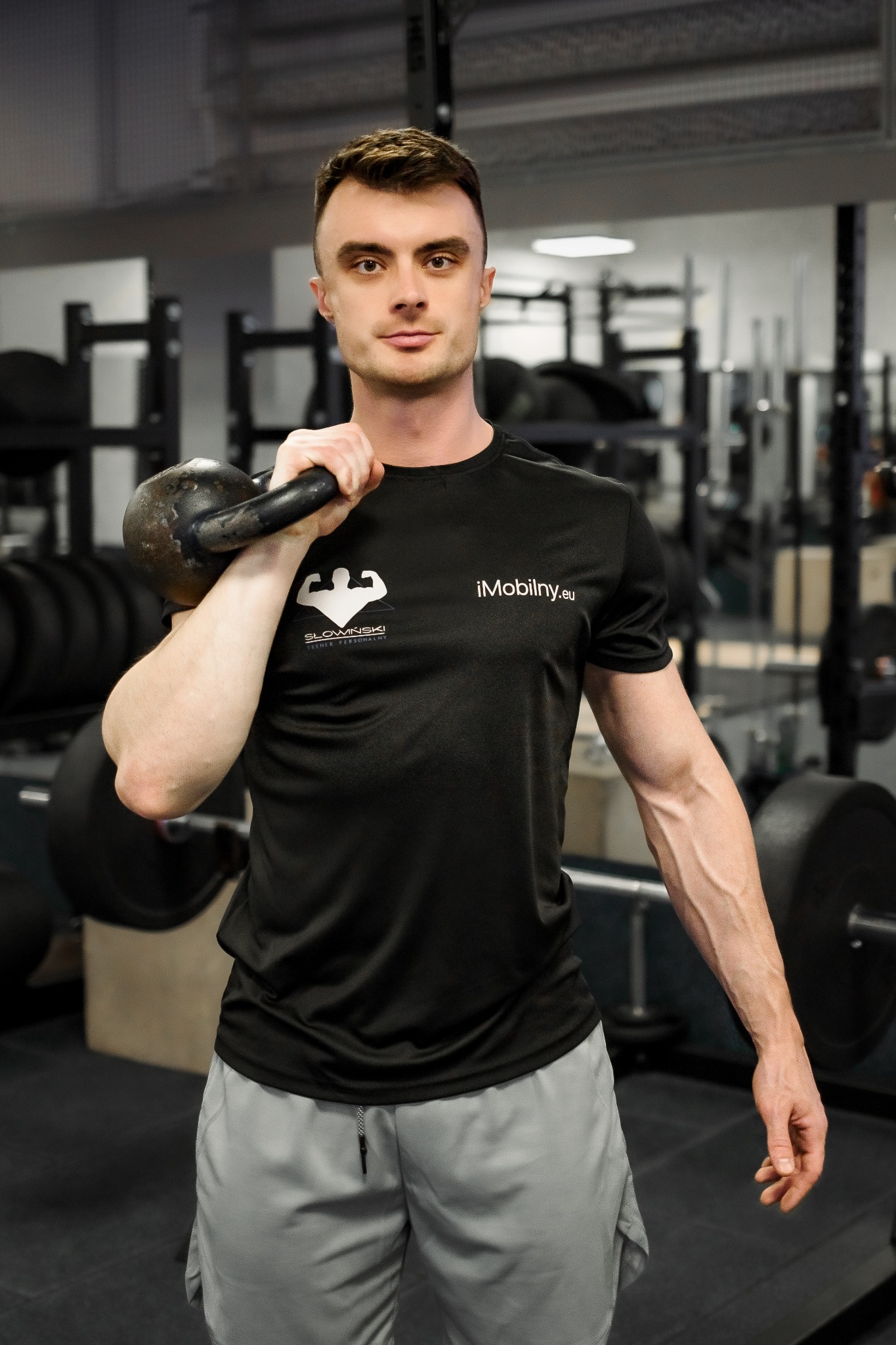 Młody mężczyzna w czarnej koszulce z logo siłowni, trzymający kettlebell na ramieniu, na tle sprzętu do ćwiczeń w siłowni.
