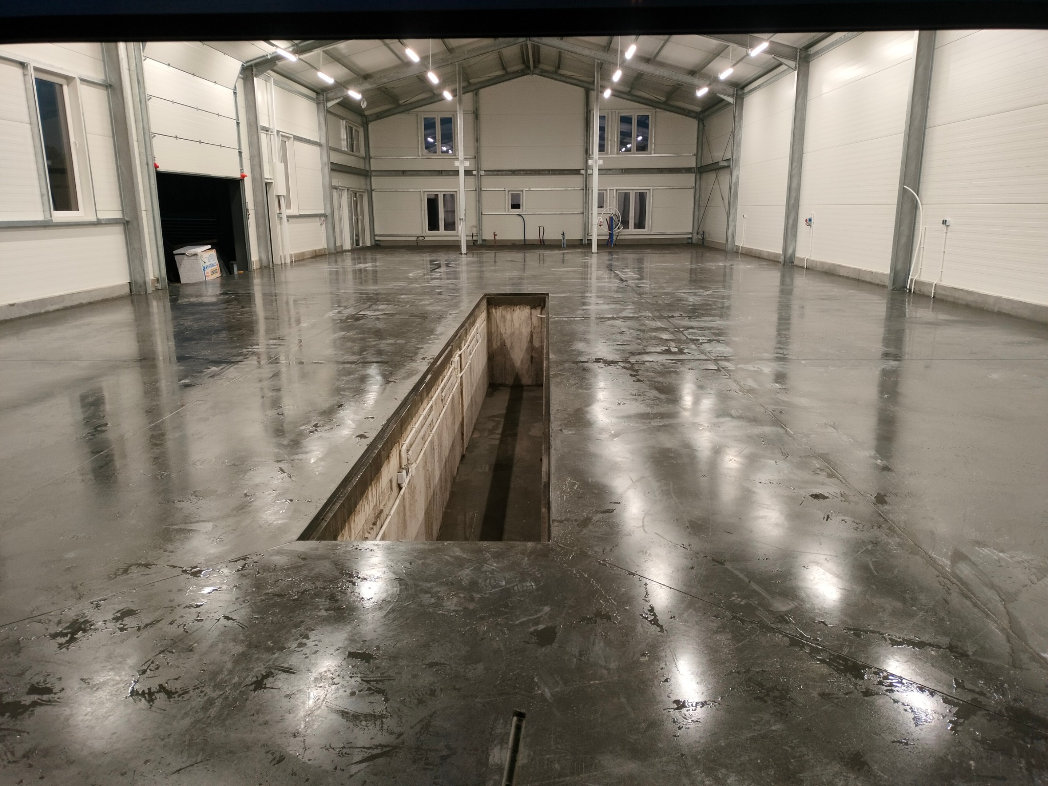 Wnętrze hali przemysłowej z szarą, błyszczącą posadzką i kanałem rewizyjnym; widoczne okna na antresoli i oświetlenie sufitowe.