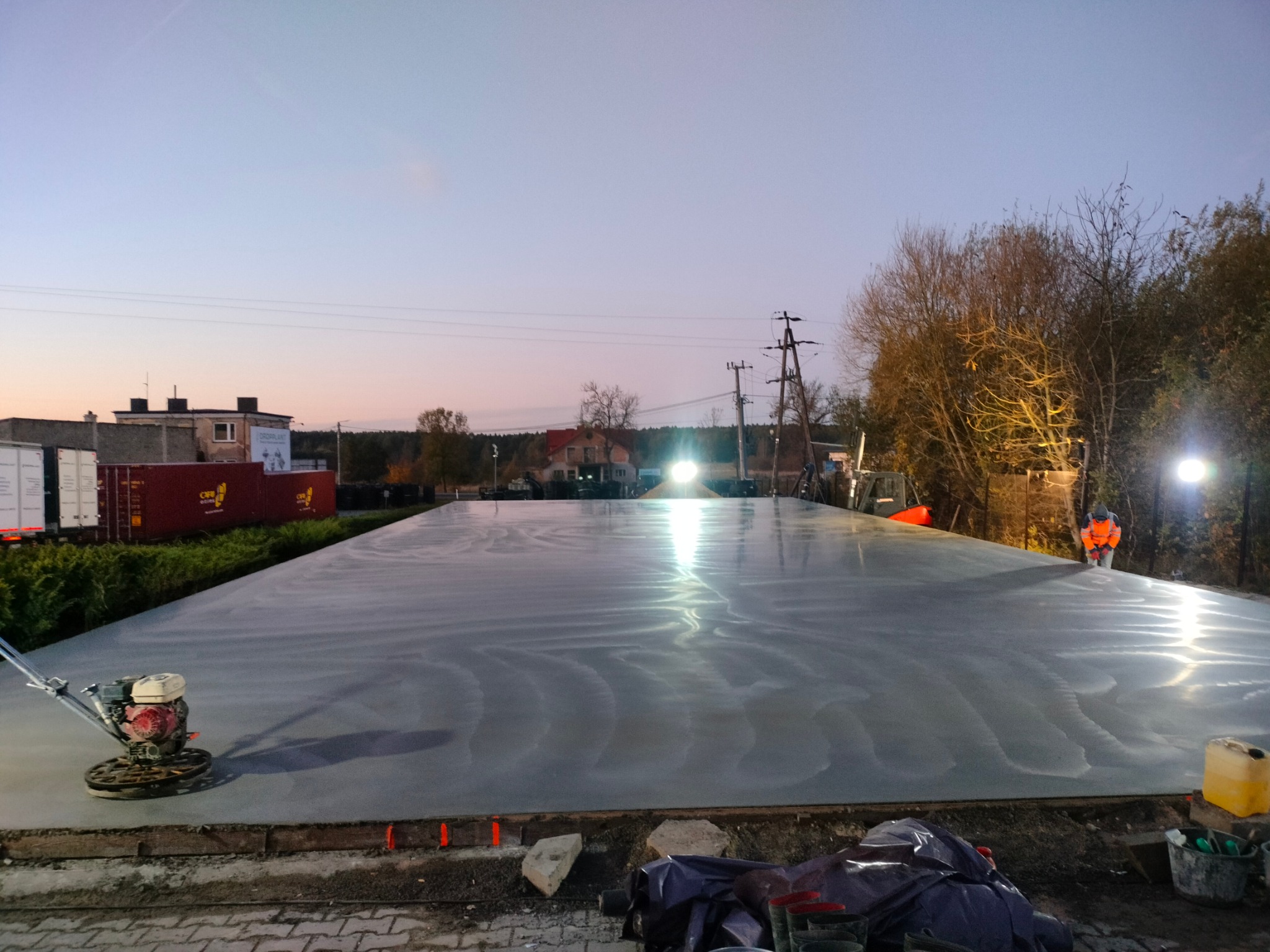 Wygładzona, mokra powierzchnia betonowa zacierana mechanicznie zacieraczką; w tle budynki i drzewa o zmierzchu.