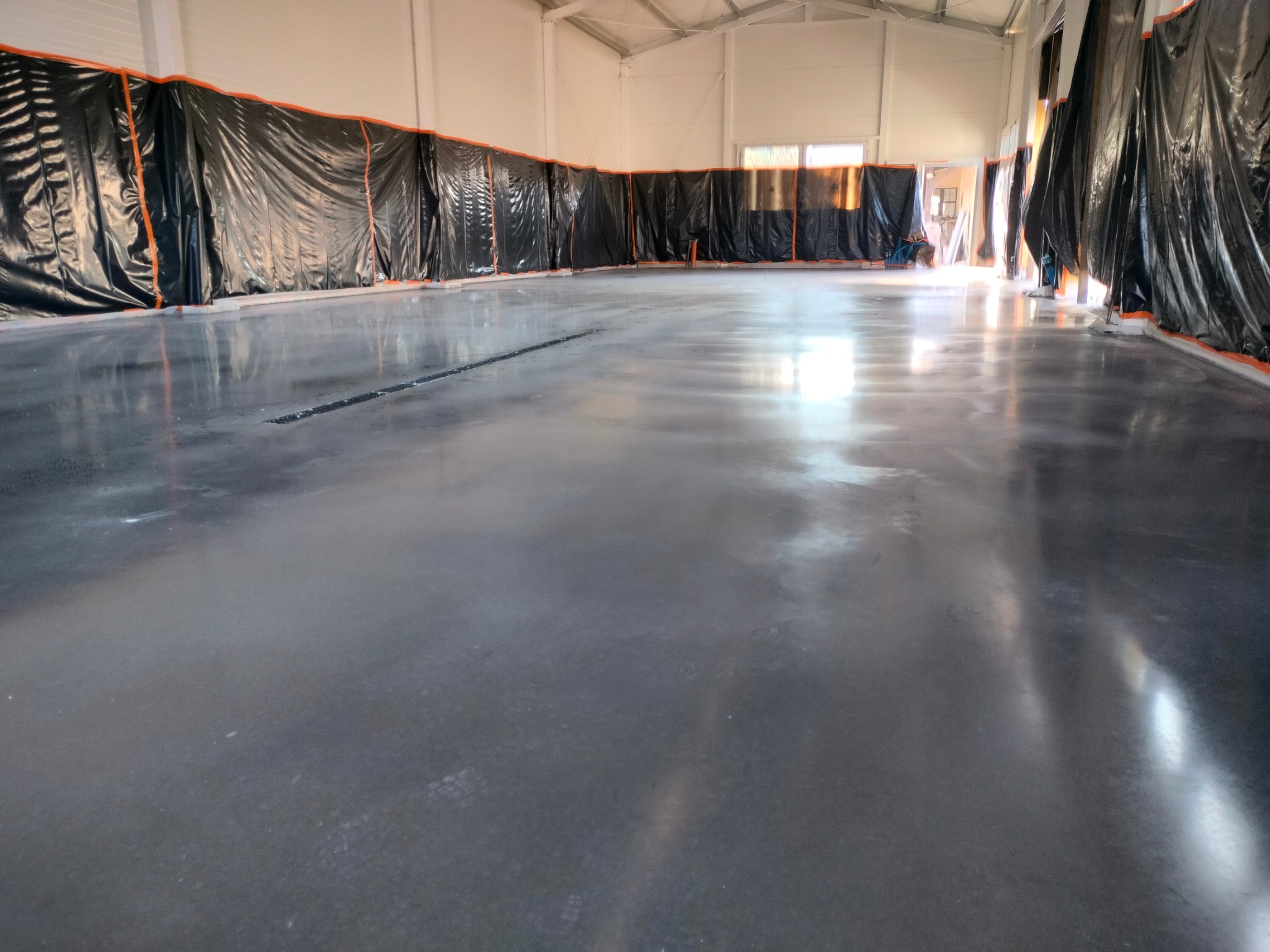 Świeżo wylana, szara posadzka w hali przemysłowej, zabezpieczona folią przy ścianach, z widocznymi śladami po narzędziach i refleksami światła.