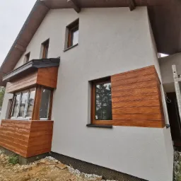 Elewacja domu jednorodzinnego z oknami w drewnianych ramach, częściowo wykończona tynkiem imitującym drewno, widoczne rusztowanie i teren wokół domu w trakcie prac wykończeniowych.