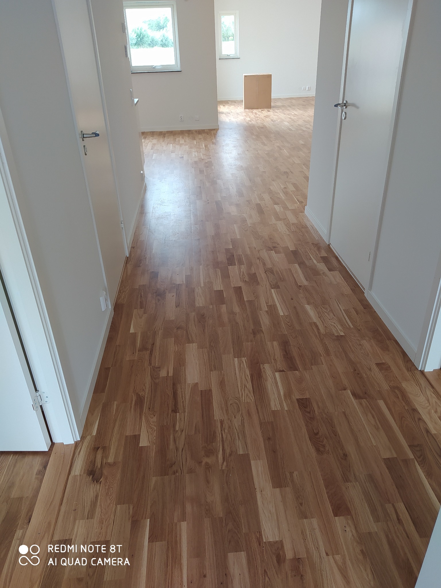 Ułożony parkiet w korytarzu z białymi drzwiami, widok na dalsze pomieszczenia z oknami, widoczny karton w tle.