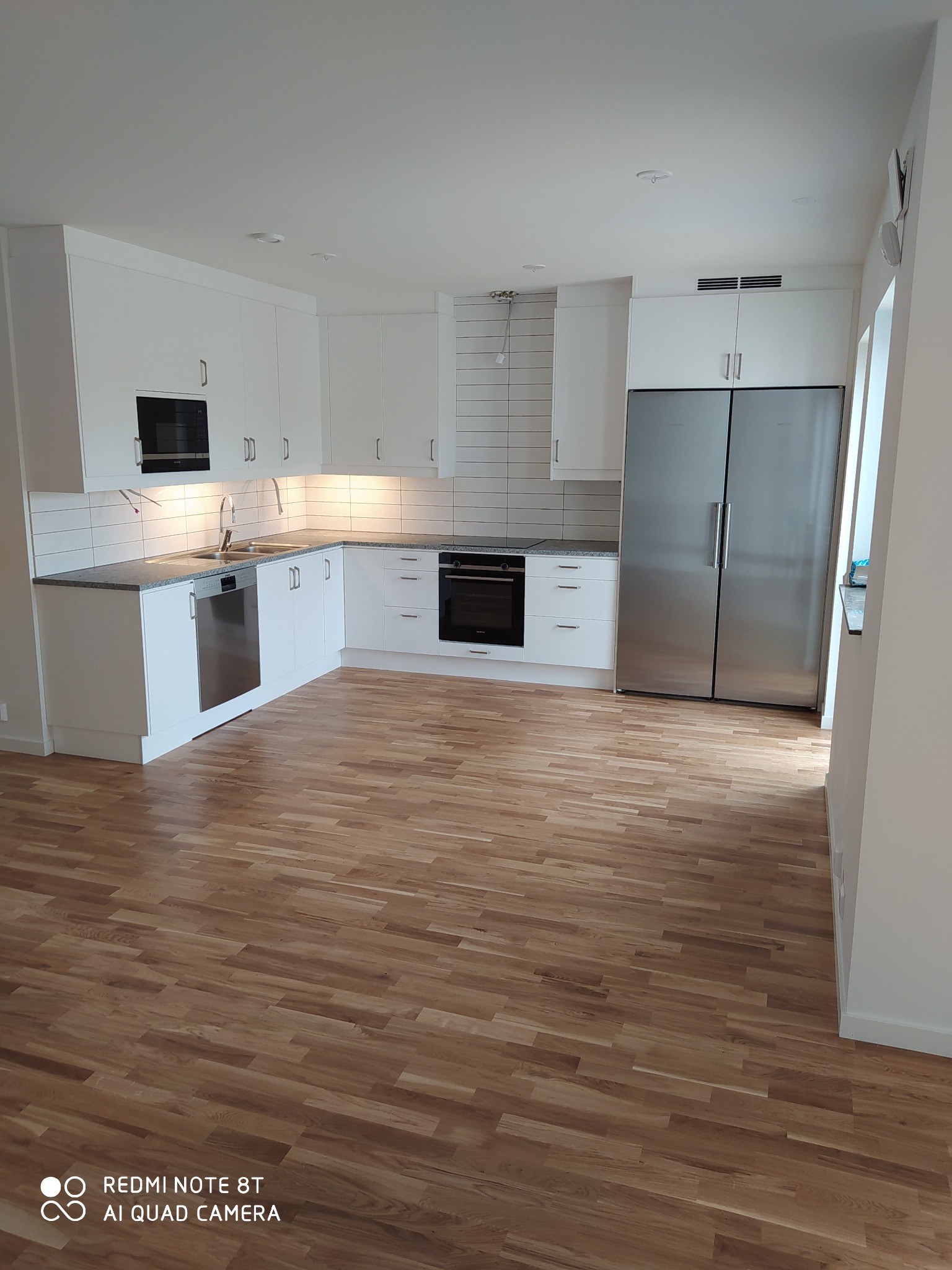 Jasna kuchnia z drewnianą podłogą, białymi szafkami, szarym blatem i sprzętem AGD ze stali nierdzewnej, widok na całe pomieszczenie.