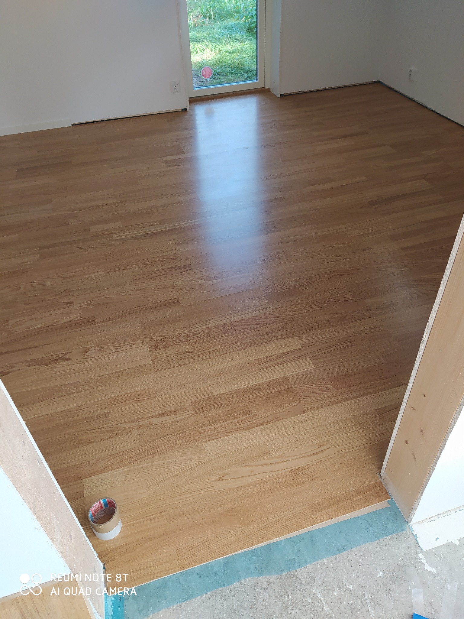 Jasna podłoga z paneli ułożona w pustym pokoju, widoczna taśma malarska przy listwie przypodłogowej i otwartych drzwiach.