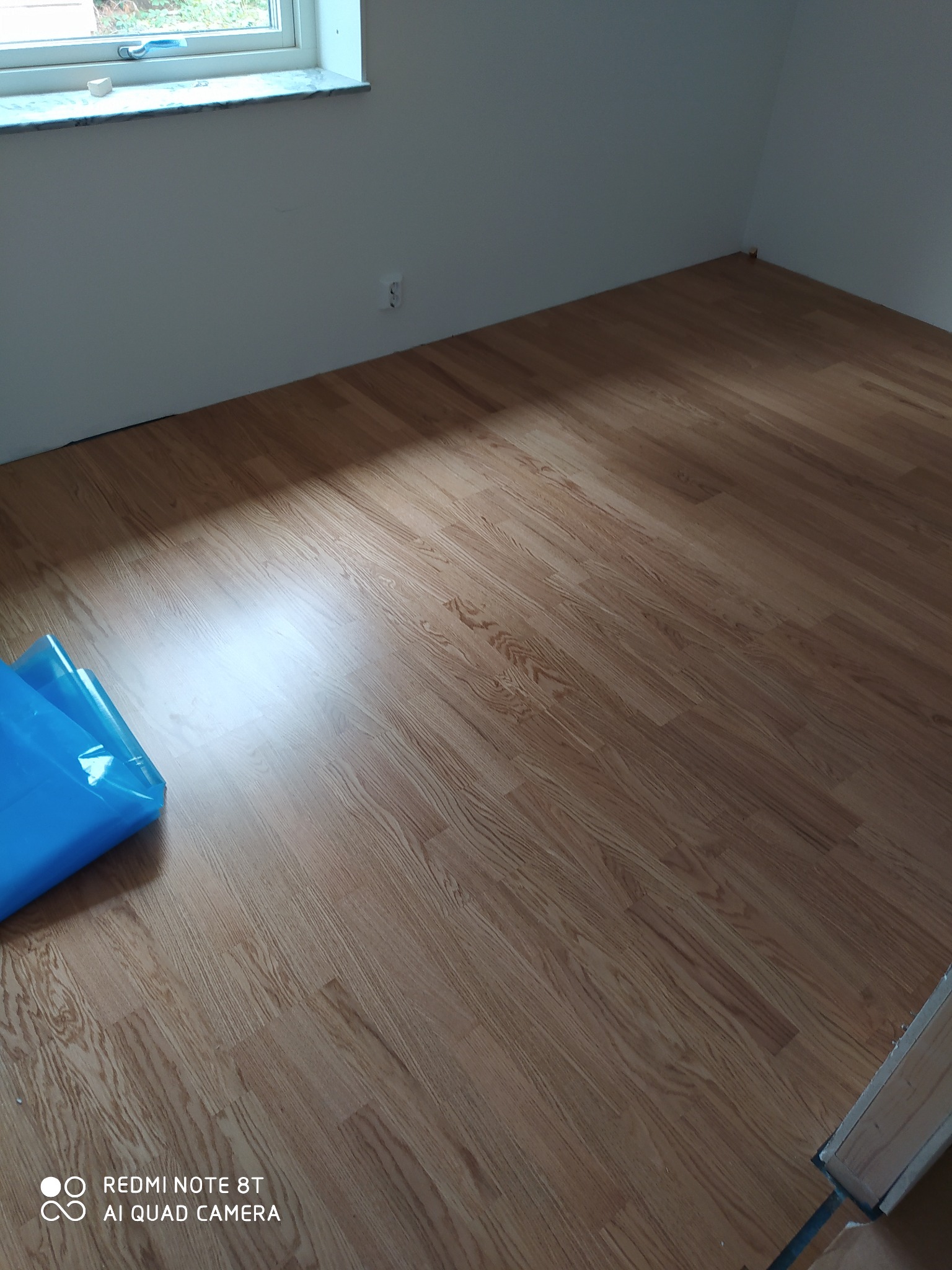 Nowo ułożone panele podłogowe o wzorze drewna w pustym pokoju, z fragmentem niebieskiej folii ochronnej w rogu.