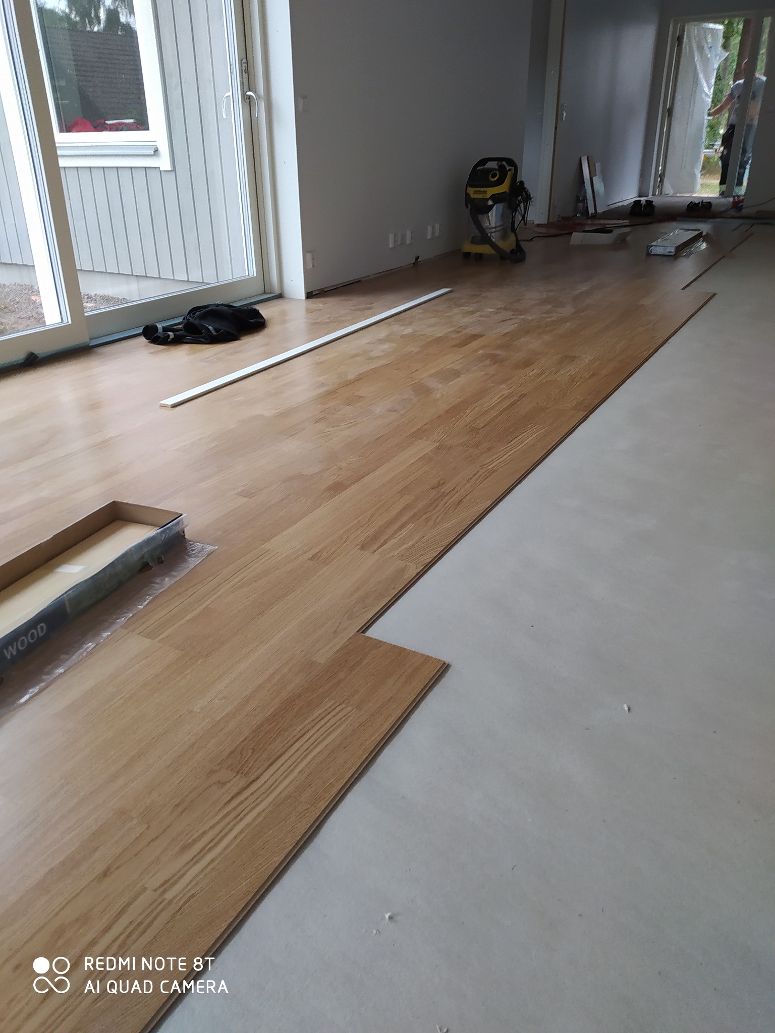 W trakcie montażu jasnych paneli podłogowych, widoczny otwarty karton z panelami, listwa przypodłogowa i odkurzacz przemysłowy w tle.