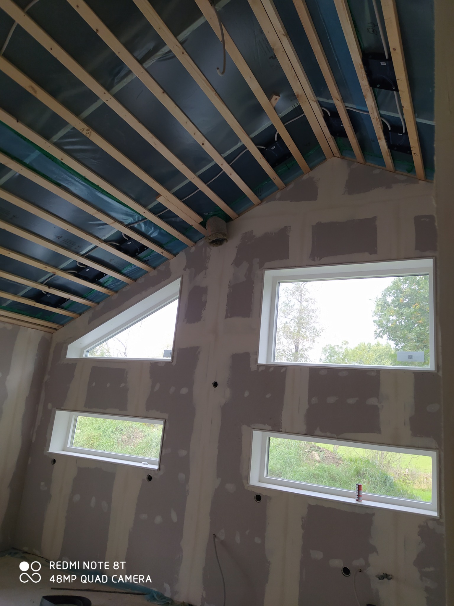 Wnętrze w trakcie prac wykończeniowych: sufit z drewnianą konstrukcją i izolacją, ściany z zamontowanymi oknami i widocznymi łączeniami płyt gipsowo-kartonowych przed szpachlowaniem.