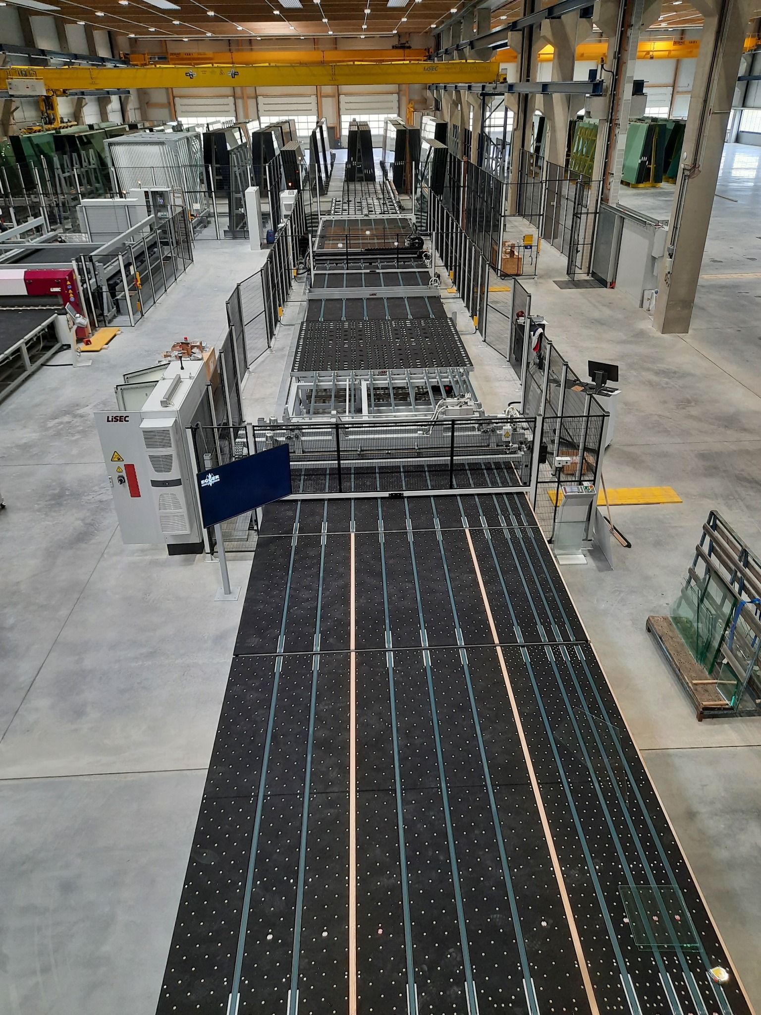 Widok z góry na linię produkcyjną w hali, z maszynami i transportem szkła, widoczne gotowe tafle szkła.