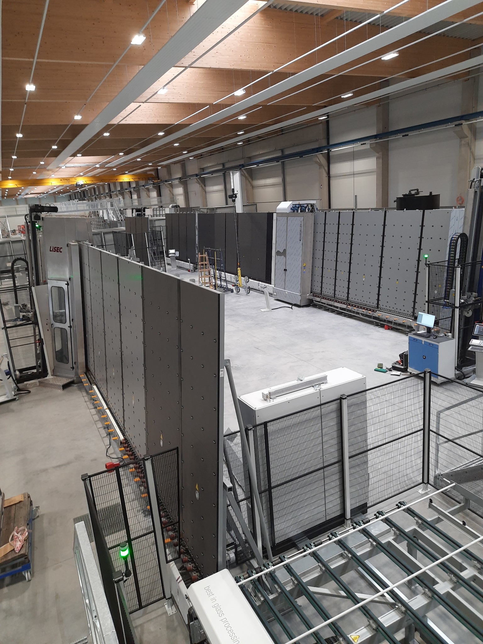 Wnętrze hali produkcyjnej z automatyczną linią do obróbki szkła, widoczna maszyna LISEC i panele szklane na rolkach transportowych.
