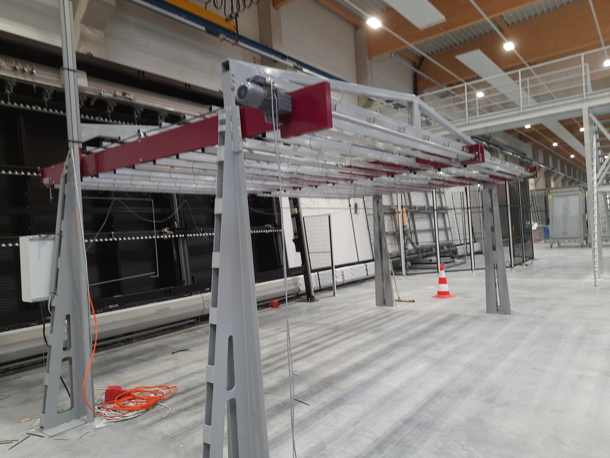 Wnętrze hali produkcyjnej z elementami konstrukcji stalowej, widoczny mechanizm suwnicy z silnikiem i czerwonymi elementami, w tle maszyny i pomarańczowy pachołek.