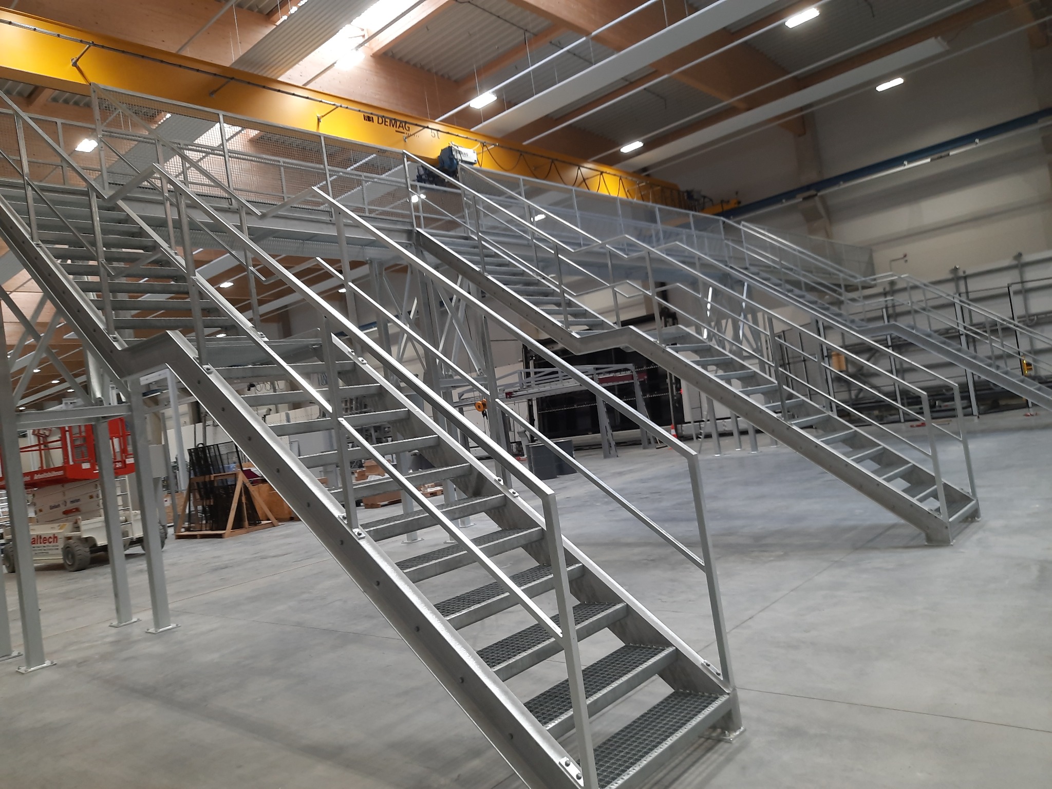 Dwie pary metalowych schodów przemysłowych z perforowanymi stopniami i barierkami, prowadzące do platformy w hali produkcyjnej, widoczny suwnica DEMAG i podnośnik Altech.