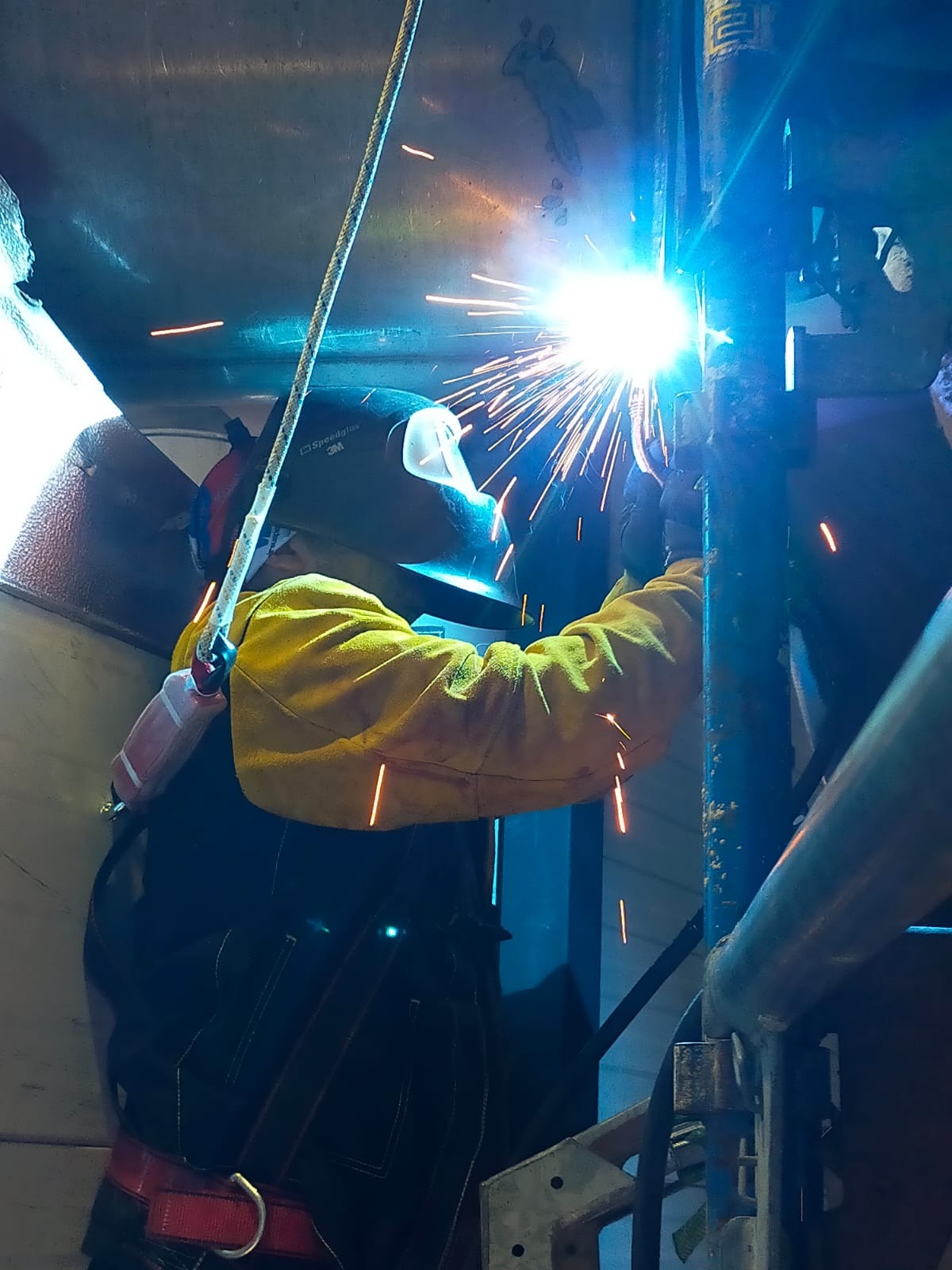 Pracownik w masce spawalniczej i żółtych rękawicach łączy metalowe elementy konstrukcji, widoczne iskry i intensywne światło procesu spawania, w tle stalowa ściana z zarysowaną postacią.