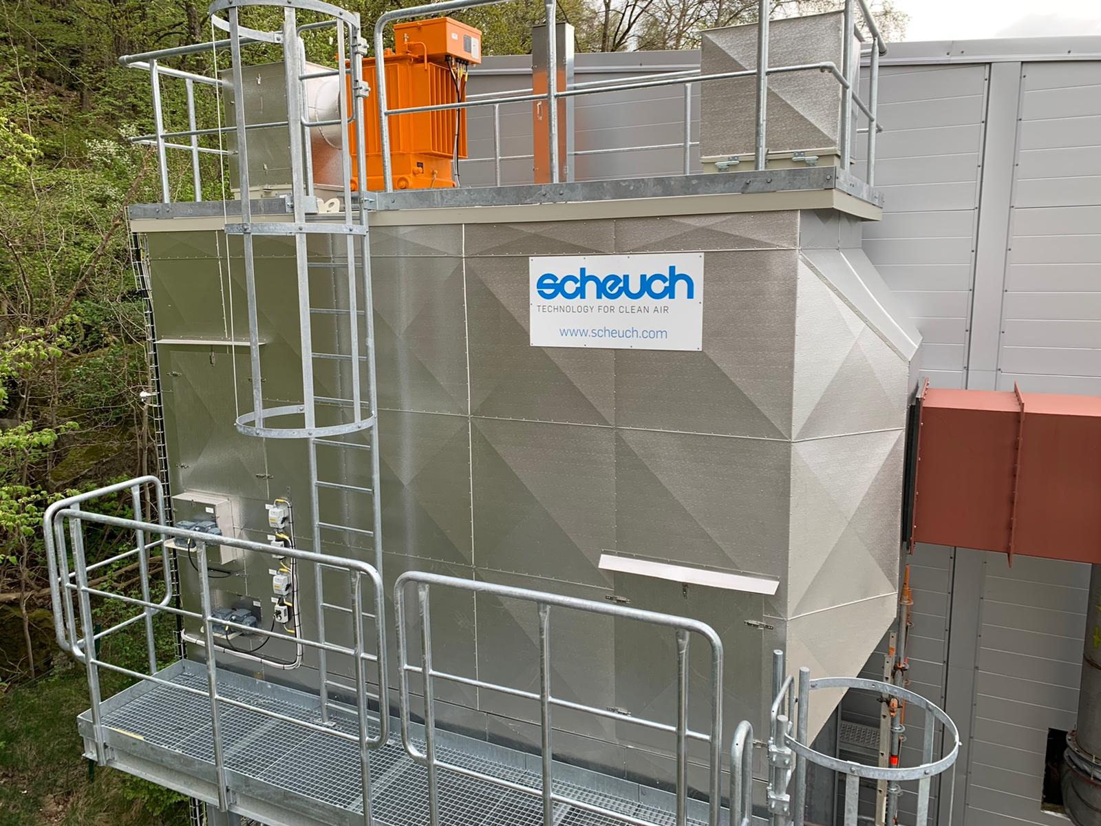 Zewnętrzna instalacja przemysłowa z metalową konstrukcją, platformą, schodami drabinkowymi i pomarańczowym silnikiem na górze, z logo firmy Scheuch promującej technologię czystego powietrza.