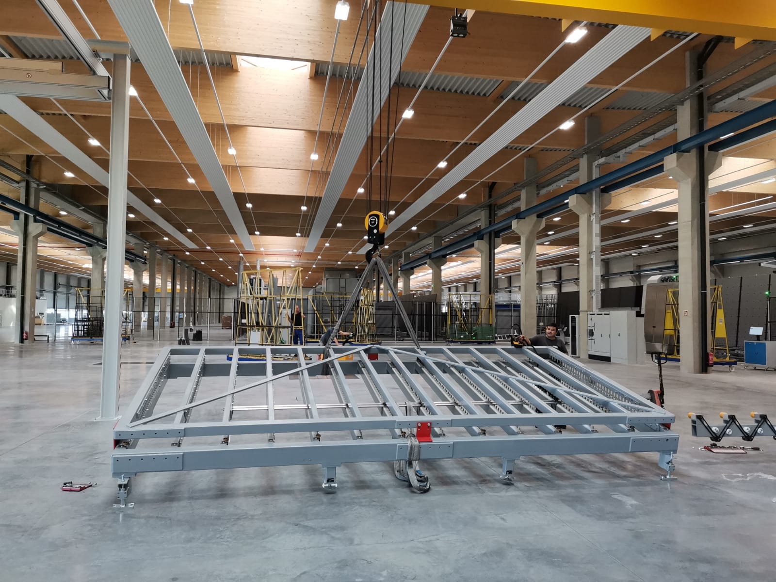 Montaż stalowej konstrukcji hali produkcyjnej z wykorzystaniem suwnicy, widoczne drewniane elementy konstrukcyjne dachu i oświetlenie przemysłowe.