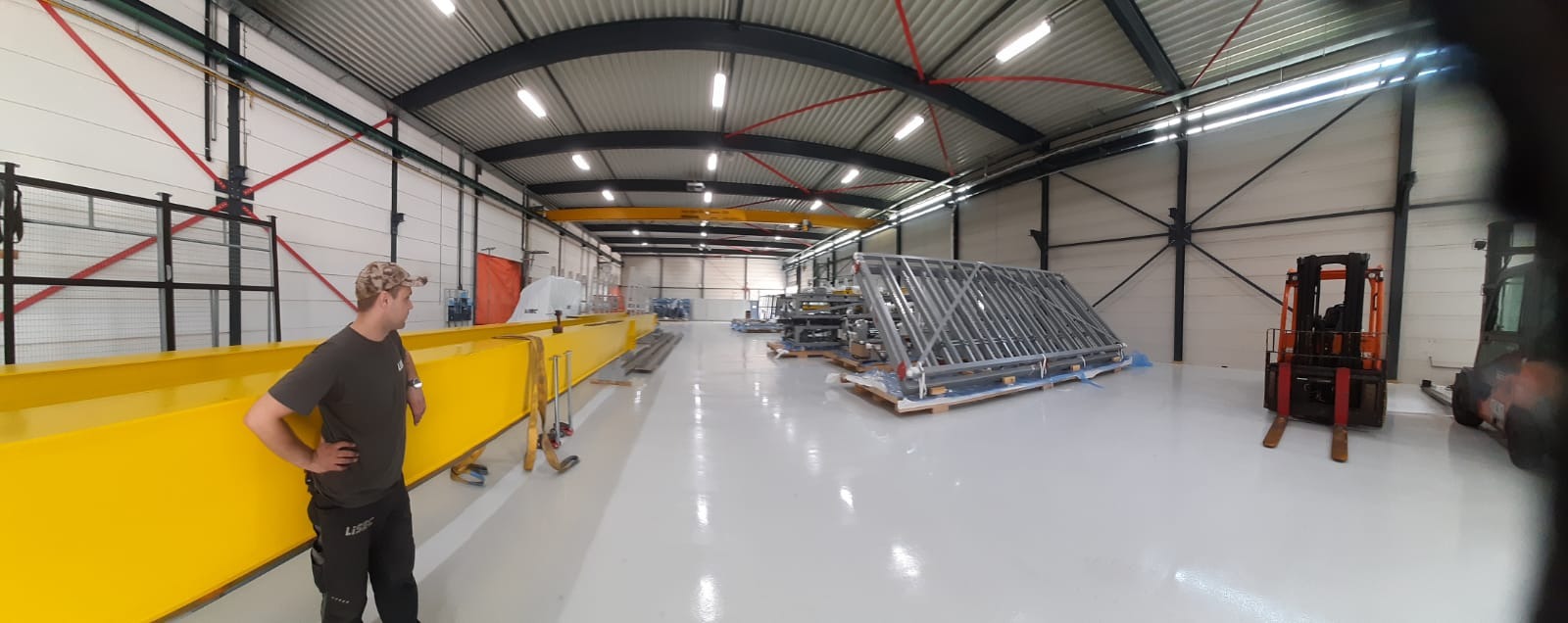 Wnętrze hali produkcyjnej z suwnicą, przygotowanymi elementami konstrukcji stalowych na paletach i wózkiem widłowym, widok z perspektywy pracownika.