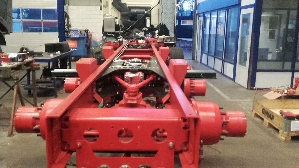 Czerwona rama podwozia ciężarówki w warsztacie, widok z przodu, przednia oś i mechanizmy napędowe widoczne, w tle brama garażowa i przeszklone biuro.