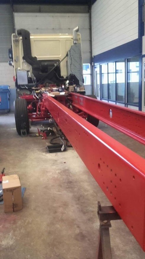 Czerwona rama ciężarówki na koziołkach w warsztacie, widoczne elementy podwozia i kabina.