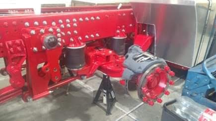 Czerwona, metalowa rama pojazdu ciężarowego z elementami zawieszenia, podparta na czarnej koziołku, widoczne elementy pneumatyczne i piasta koła z nakrętkami.