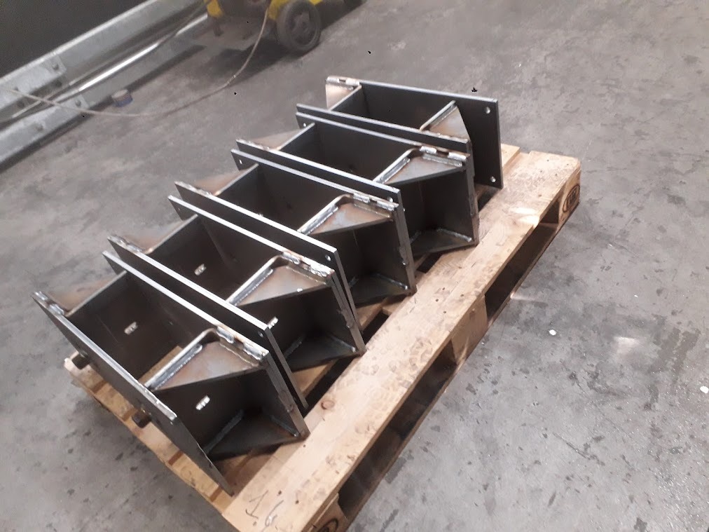 Surowe, spawane elementy konstrukcyjne z metalu, ułożone na drewnianej palecie w hali produkcyjnej, widoczne ślady spawania.