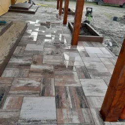 Świeżo ułożona, mokra kostka betonowa w odcieniach brązu i szarości, tworząca ścieżkę z drewnianymi słupami pod zadaszeniem, z widocznymi elementami przygotowania podłoża i narzędziem do cięcia...