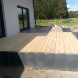 Drewniany taras obudowany szarą kostką, przylegający do jasnego budynku z dużymi oknami, w tle widoczny trawnik i las.