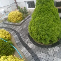 Ścieżka z kostki brukowej w odcieniach szarości i czerni, wijąca się między rabatami z ozdobnymi krzewami, prowadząca do schodów kamiennych.