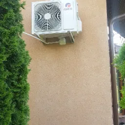 Biała jednostka zewnętrzna klimatyzatora marki Gree zamontowana na beżowej elewacji domu, widoczna instalacja odprowadzająca skropliny, obok zielona tuja.