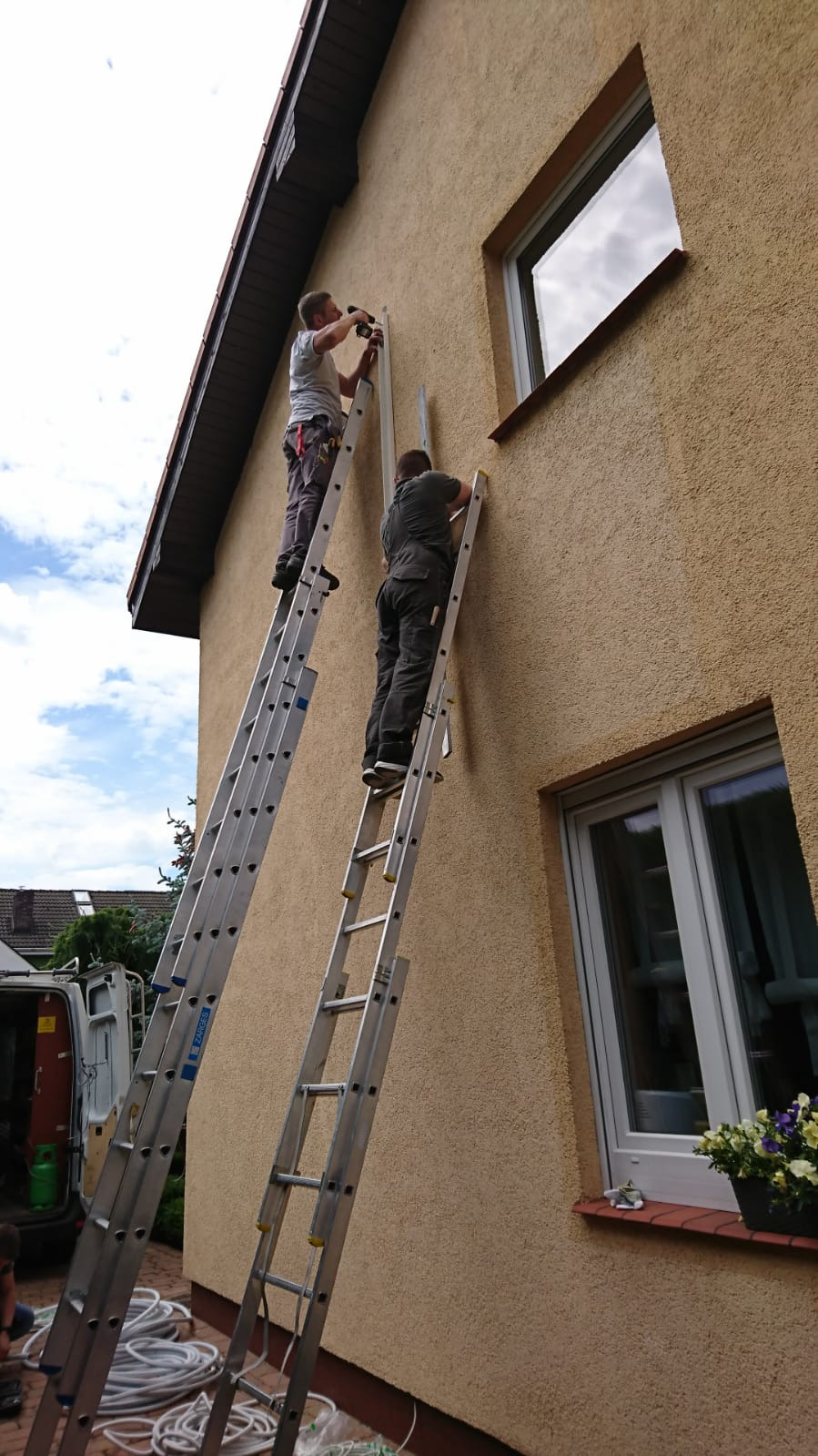 Dwóch monterów na drabinach instaluje element na elewacji domu o beżowej, chropowatej strukturze; widoczne okna i zwinięte kable na ziemi.