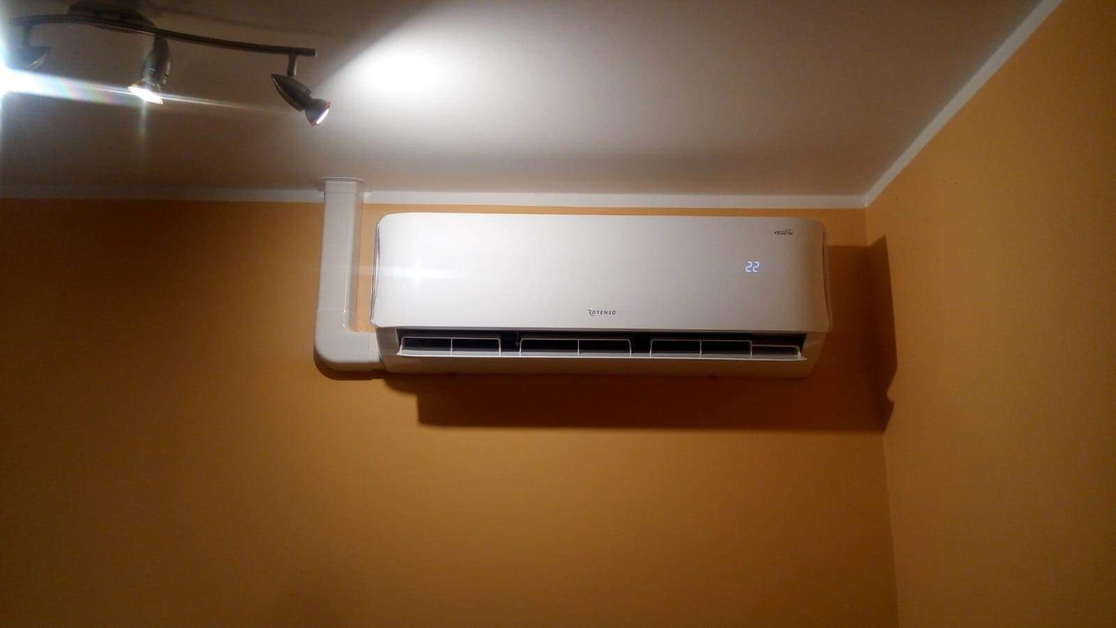 Zamontowana biała klimatyzacja Rotenso na pomarańczowej ścianie, widoczne rury odprowadzające, oświetlenie halogenowe sufitowe.