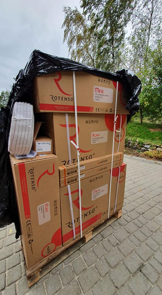 Paleta z kartonami klimatyzatorów Rotenso i Ivensis, zabezpieczona czarną folią, na kostce brukowej.